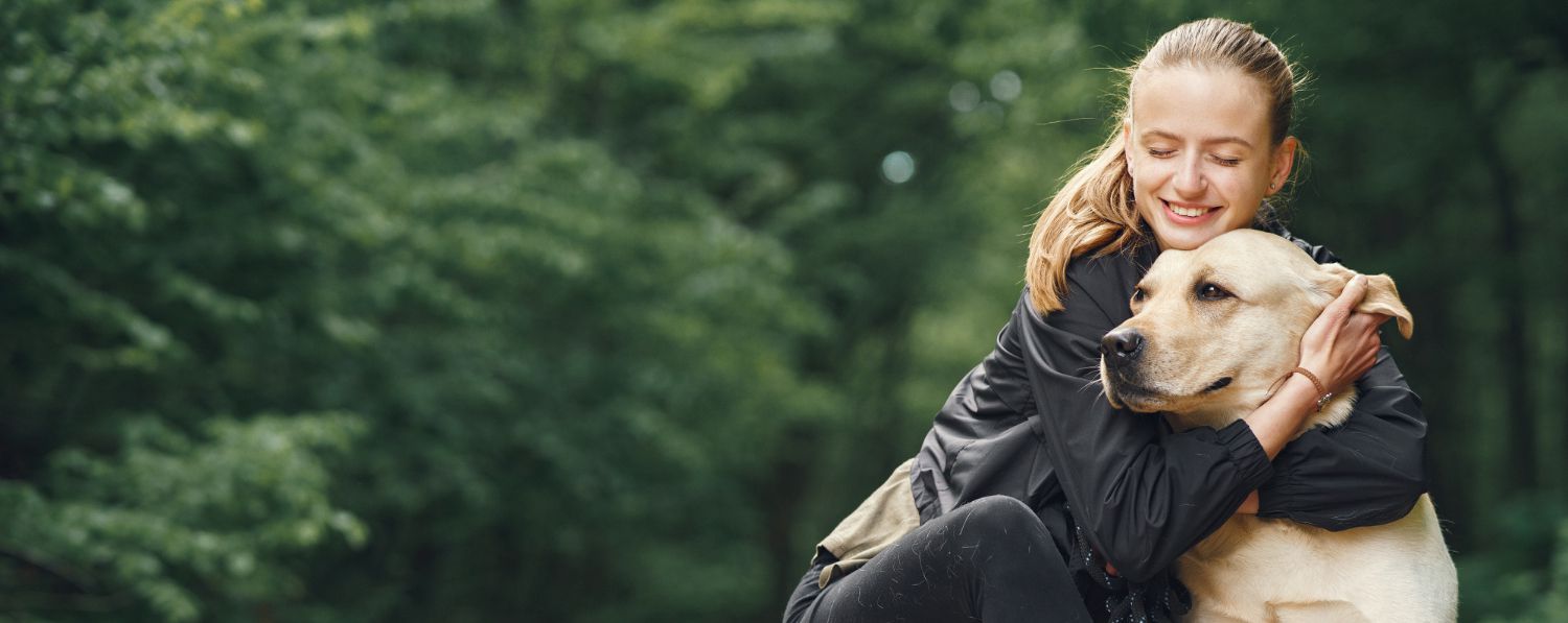 Egy fiatal nő szeretettel átöleli kutyáját az erdőben, ami a kutyatartás és nevelés harmóniáját fejezi ki.